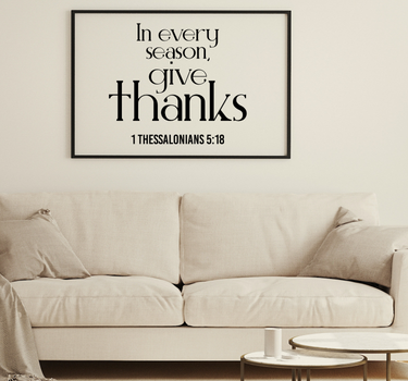 A framed wall art print with the quote 'In every season, give thanks 1 Thessalonians 5:18' in black text on a white background, hanging above a light beige sofa in a styled living room.